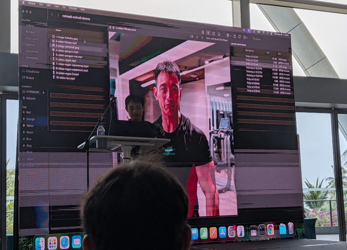 Speaker at a lectern demonstrating a video on a large screen
