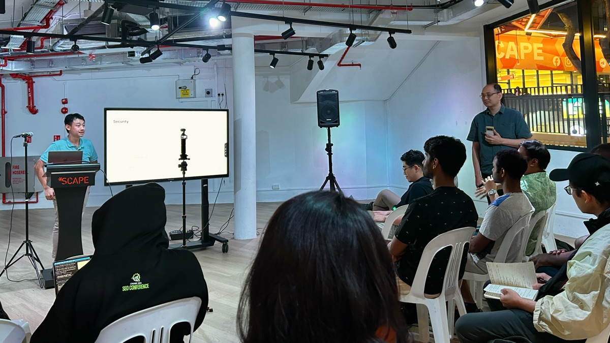 Speaker at a lectern beside a security slide with attendees seated nearby